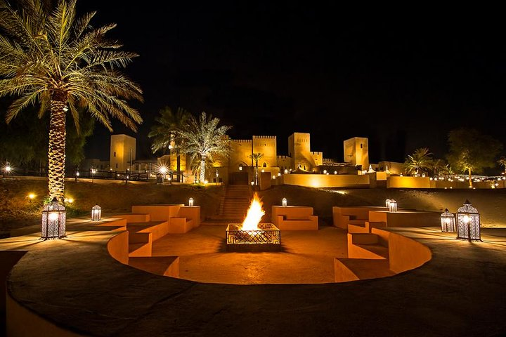 Experience the magic of Arabian nights with warm fires traditional seating and exquisite cuisine nestled among palm trees and elegant structures under the starry desert sky.